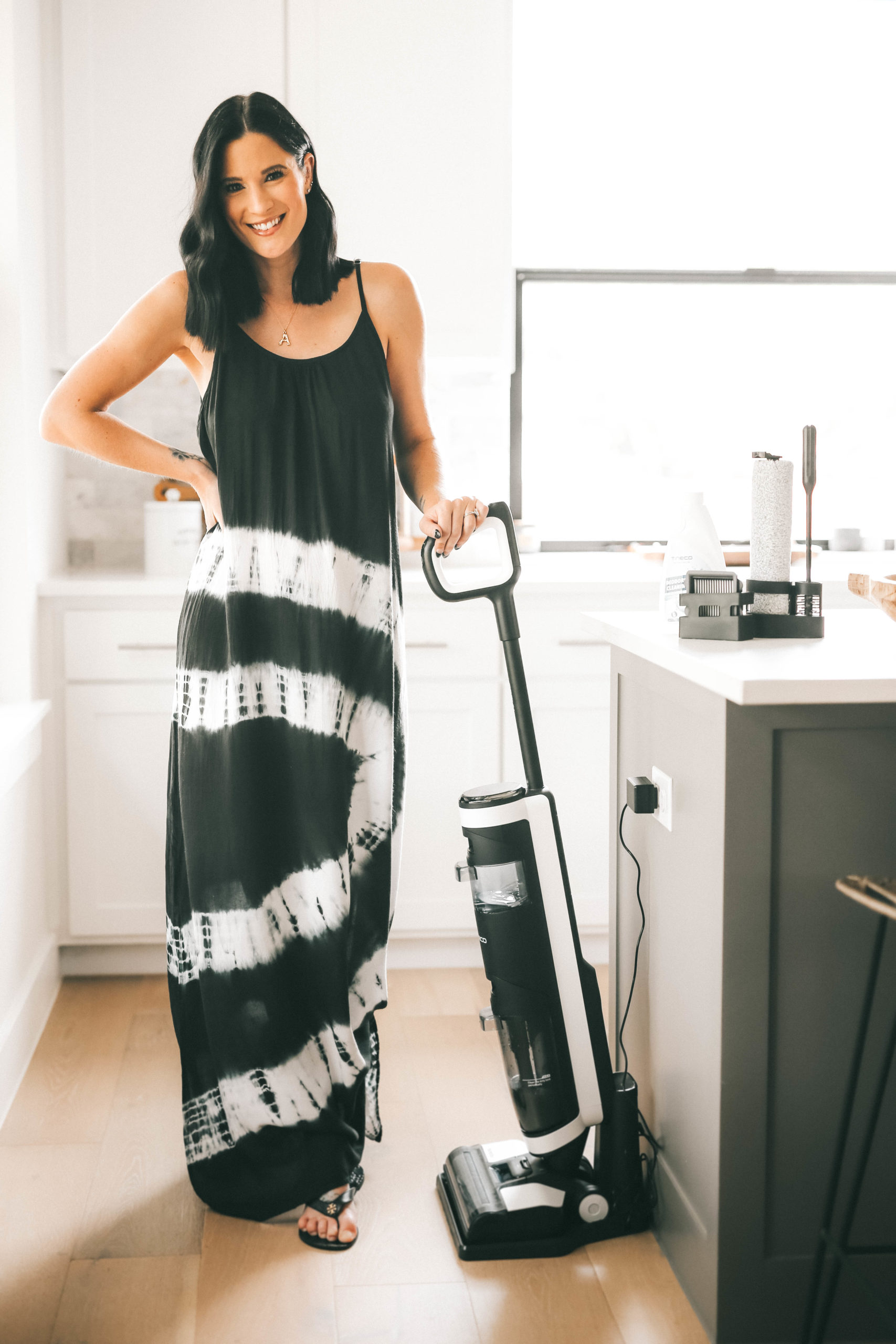 Tineco Floor One S3 by popular Austin lifestyle blog, Dressed to Kill: image of a woman wearing a black and white tie dye maxi and standing in her kitchen next to a Tineco Floor One S3 vacuum.