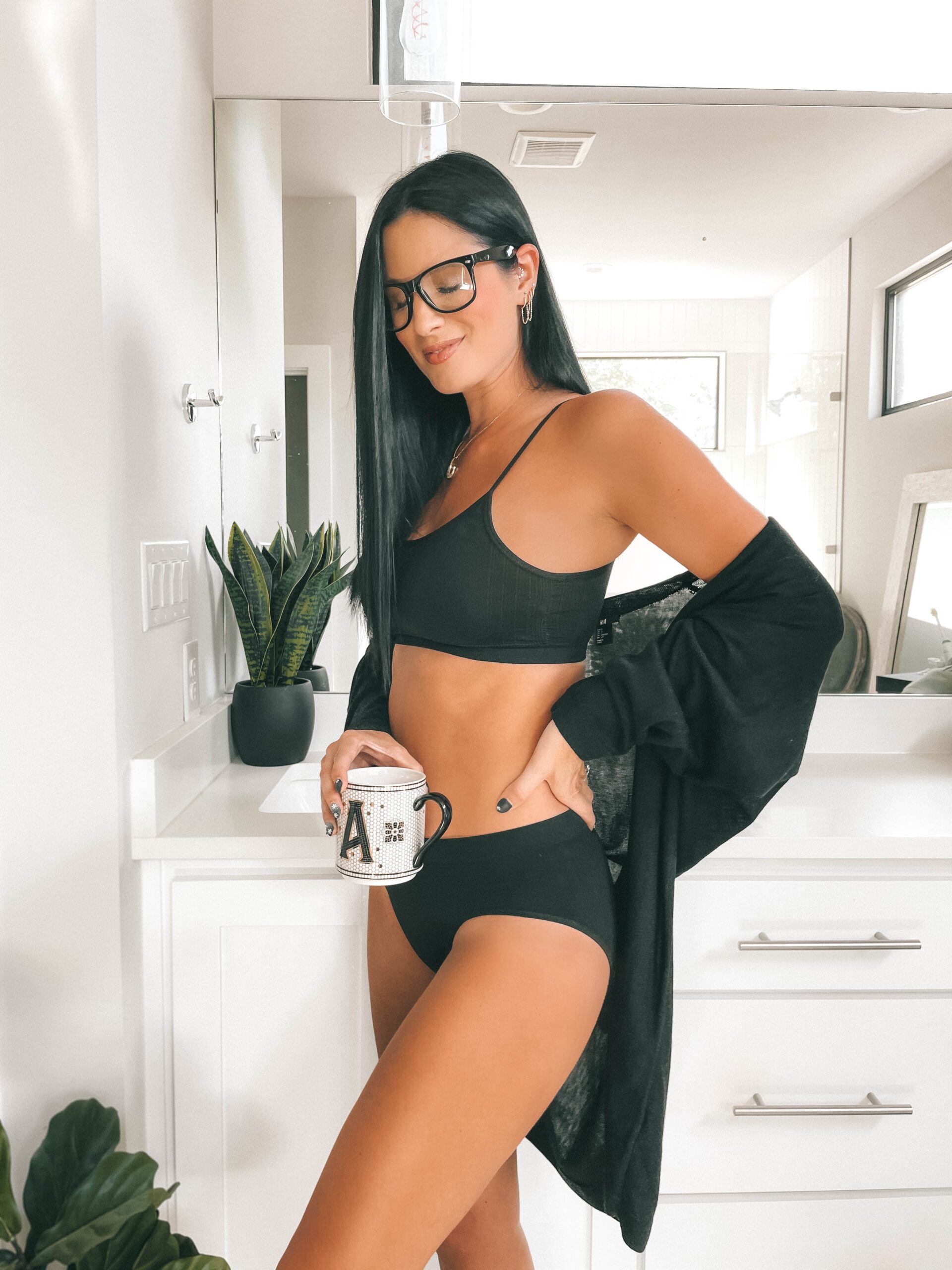 Comfy Bras by popular Austin fashion blog, Dressed to kill: image of a woman standing in her bathroom and holding a monogram mug while wearing a black duster sweater, black frame glasses, and black bra and undies set. 